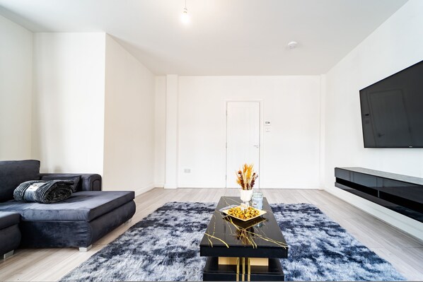 A modern living room with a black sofa, a black coffee table with a vase, and a flat-screen TV mounted on the wall. - LUX 2bdr Flat Near LTN Airport City Centre (Luton)