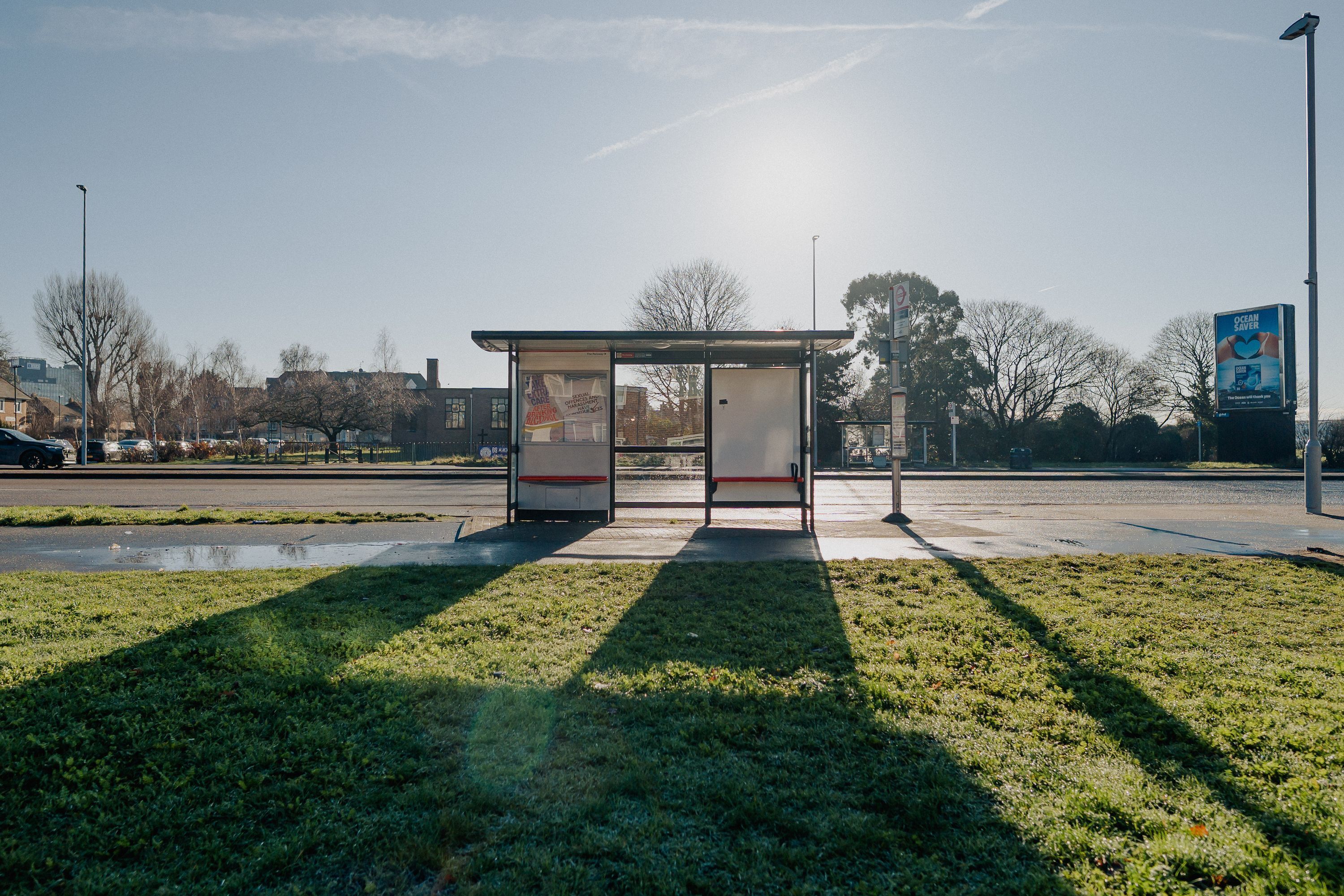 Un refugio de autobús con techo transparente, situado en un área cubierta de césped con sombras proyectadas sobre el suelo.