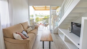Una sala moderna con un sofá, una mesa de centro, un televisor sobre un estante blanco y un balcón con mobiliario exterior.