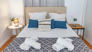 Una cama bien hecha con ropa de cama blanca y azul, un plaid estampado y dos toallas en el suelo.