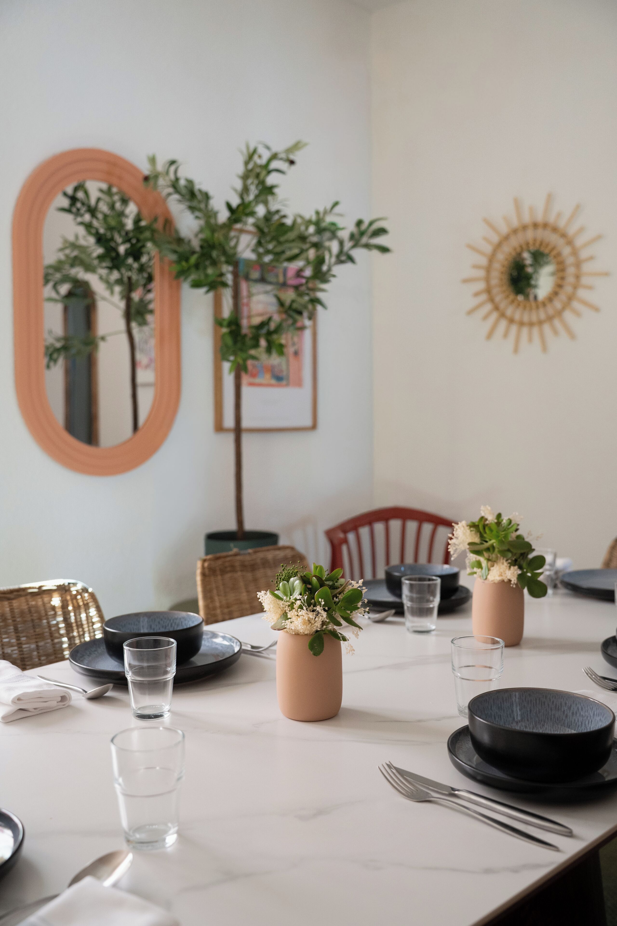 Une table à manger dressée avec des assiettes, des bols, des verres et des couverts, entourée de chaises en osier et de plantes en pot.