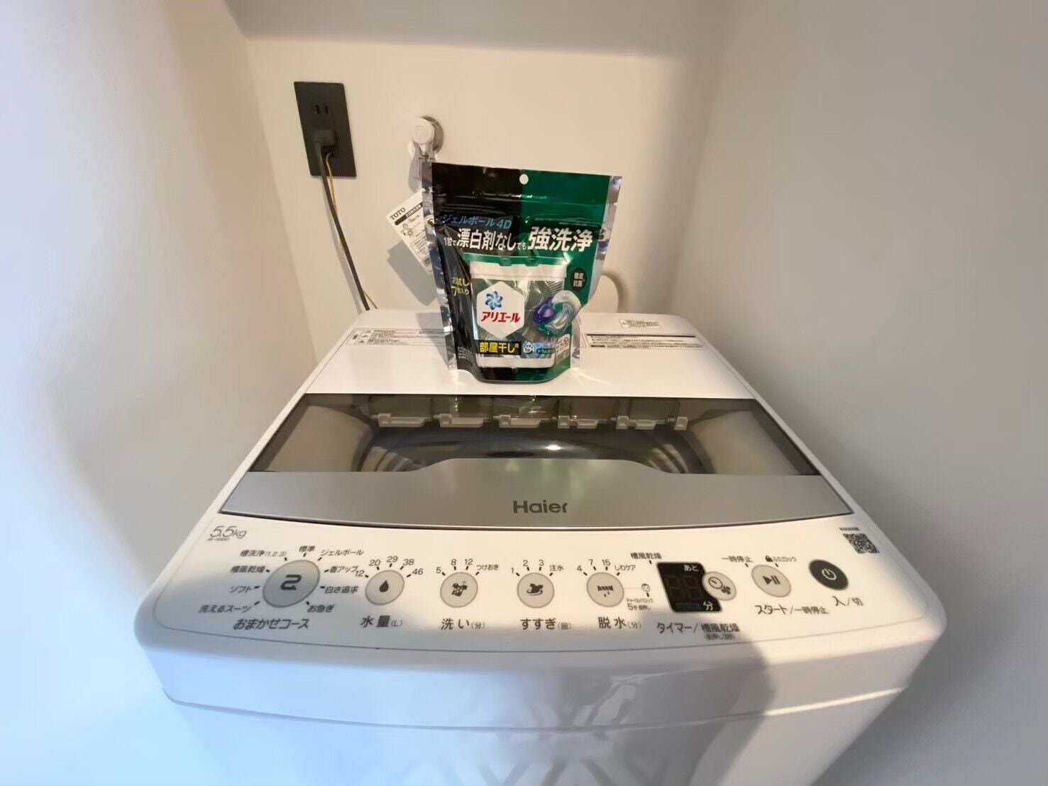 Una lavadora Haier blanca con puerta transparente, un panel de control con instrucciones en japonés y una bolsa de detergente verde encima.