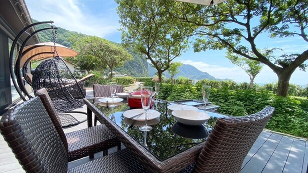 An outdoor dining area with wicker furniture, a glass table, and a view of the landscape. - Seaside Garden Nishiizu 1  (Nishiizu)