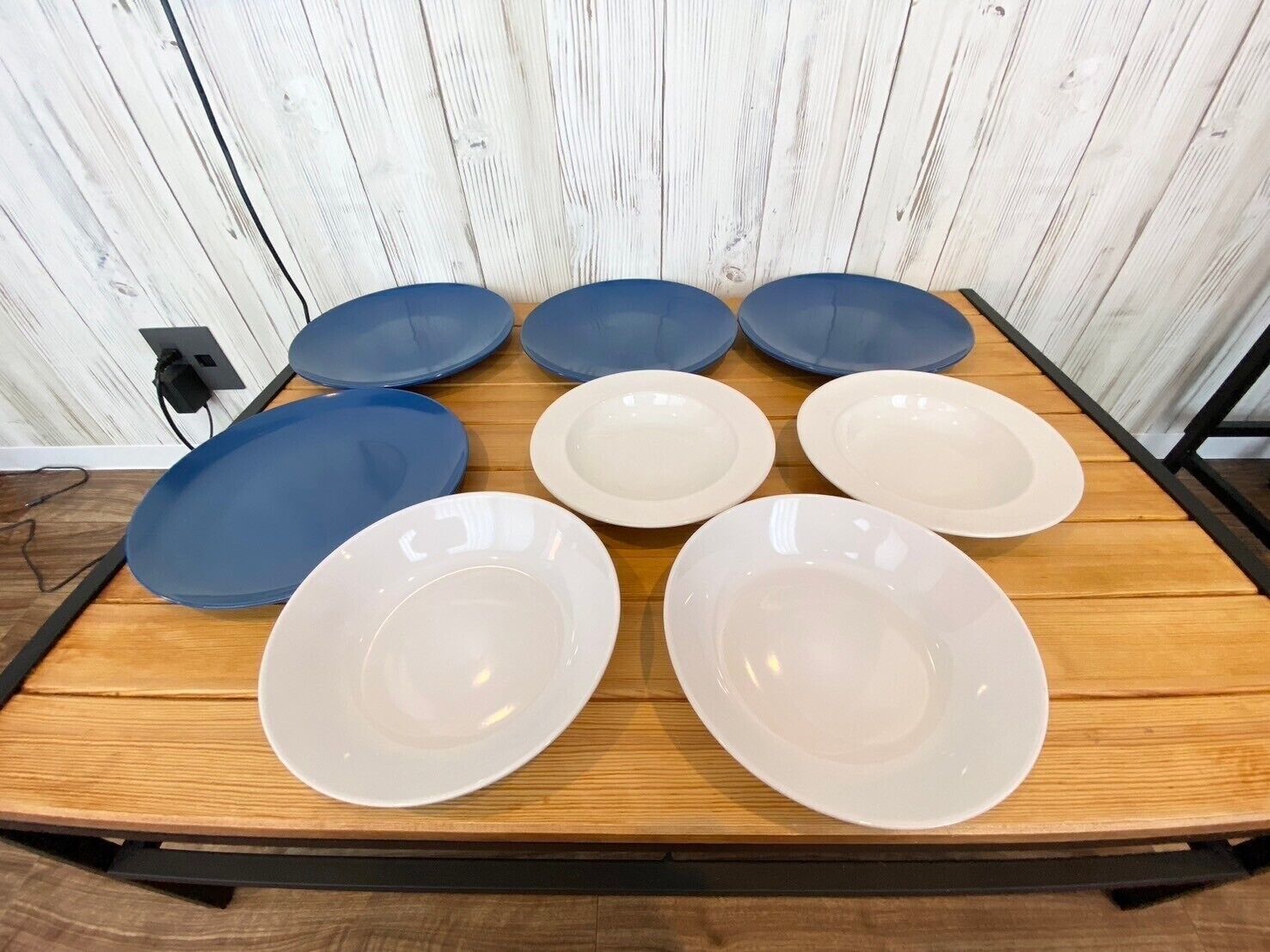 A table with six plates, three blue and three white, arranged on a wooden table against a white wooden backdrop.