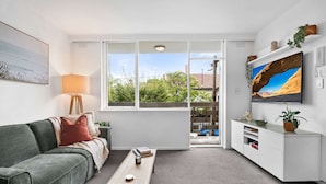 A living room with a sofa, coffee table, TV, and a window with a view of a car and greenery outside. - St Kilda Retreat Near Beach & Cafes (Melbourne)