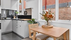 A modern kitchen with white cabinetry, a wooden dining table, and a window with a view of a brick building. - St Kilda Retreat Near Beach & Cafes (Melbourne)
