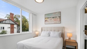 A bedroom with a large bed, two bedside tables with lamps, a painting on the wall, and a window with a view of a house and trees. - St Kilda Retreat Near Beach & Cafes (Melbourne)