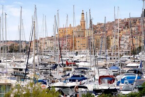 Un port de plaisance où de nombreux bateaux sont amarrés, un bâtiment imposant avec une tour en arrière-plan, et une zone urbaine densément peuplée.