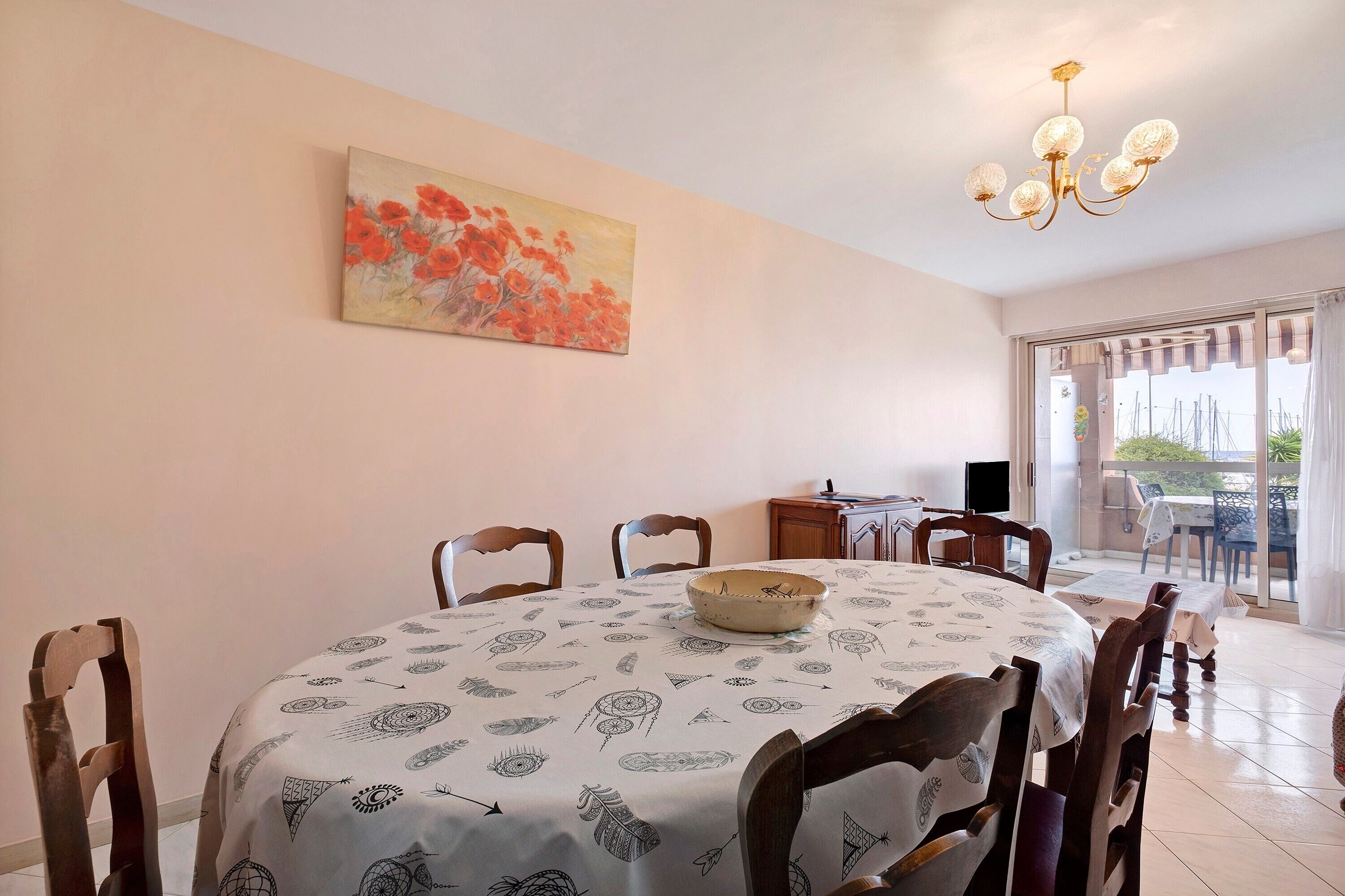 A dining area with a round table, chairs, a chandelier, a painting, and a view through a glass door.