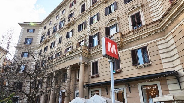 A multi-story building with a prominent 'M' sign, storefronts, and a tree in the foreground. - V10 Guest House - Vanvitelli 10 (Naples)