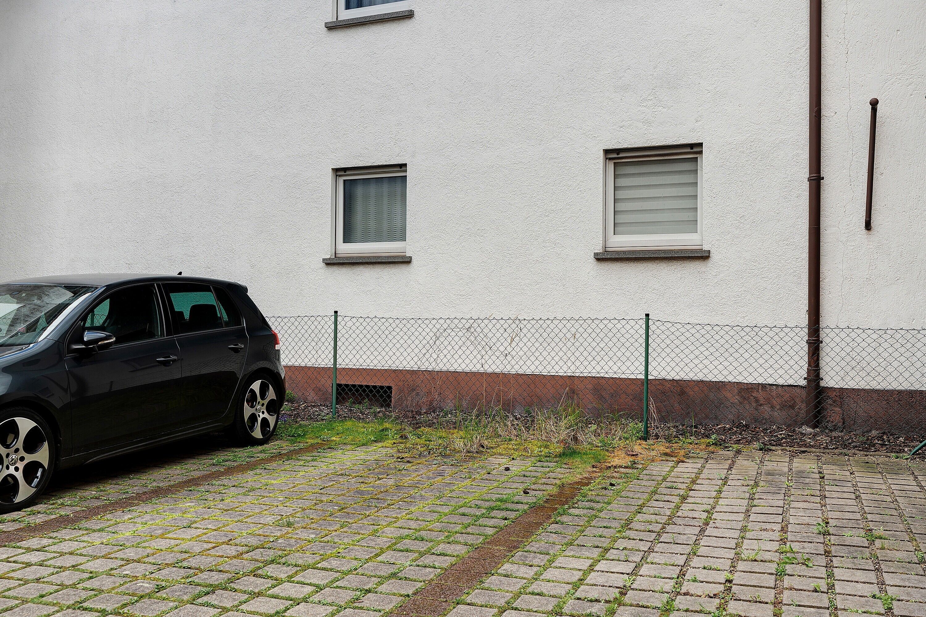 A car parked in front of a building with a chain-link fence, a paved area with grass patches, and a visible window.