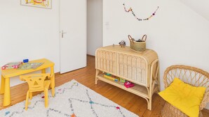 Une pièce avec un plancher en bois, une porte blanche, une table jaune avec une chaise, une petite table d’appoint en osier avec un panier, une chaise en osier avec un coussin jaune et un tapis blanc à motifs colorés.