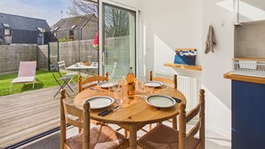 Un coin repas avec une table dressée pour quatre personnes, un espace cuisine doté d’un plan de travail bleu et une vue sur le jardin arrière.