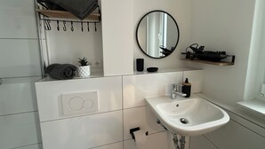 Een badkamer met een witte wasbak, een spiegel, een handdoekhouder, een plank met een handdoek, een kleine potplant en een zeepdispenser.