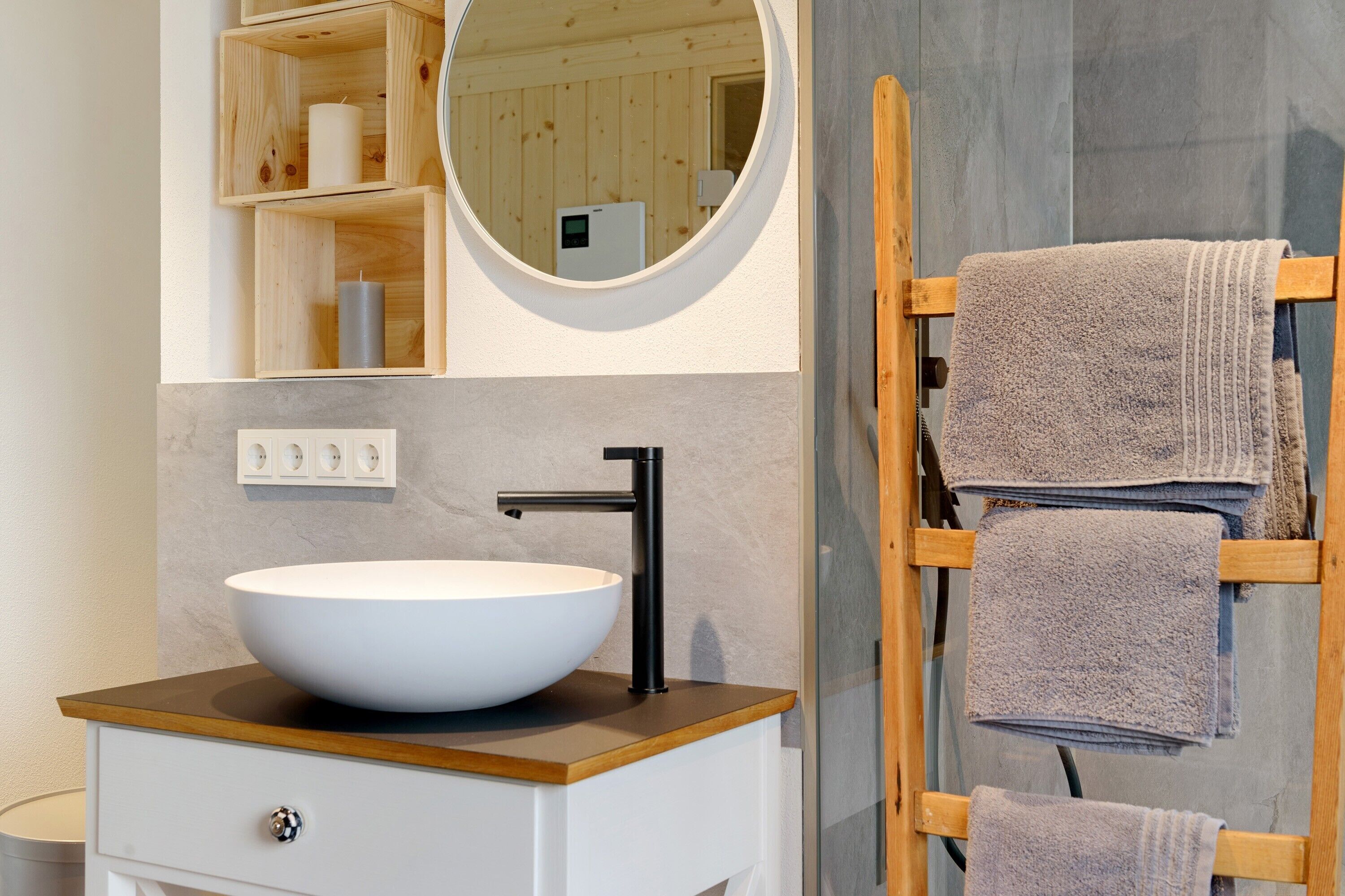 Un baño con un espejo redondo grande, un lavabo blanco tipo cuenco, repisas de madera y un perchero para toallas.