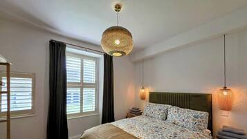A bedroom with a bed, a hanging light fixture, a window with curtains, and a bedside table.
