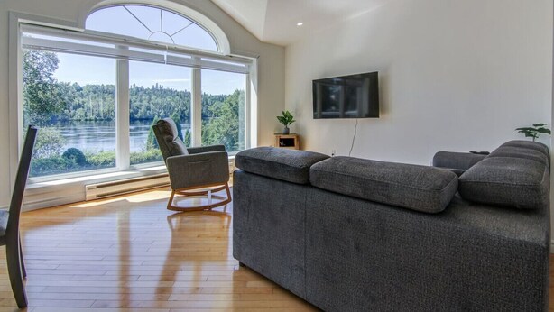 A modern living room with a large window, a grey sofa, a rocking chair, and a flat-screen TV. - L`échappée du Lac-Saint-Jean (Lac-Saint-Jean-Est Regional County Municipality)