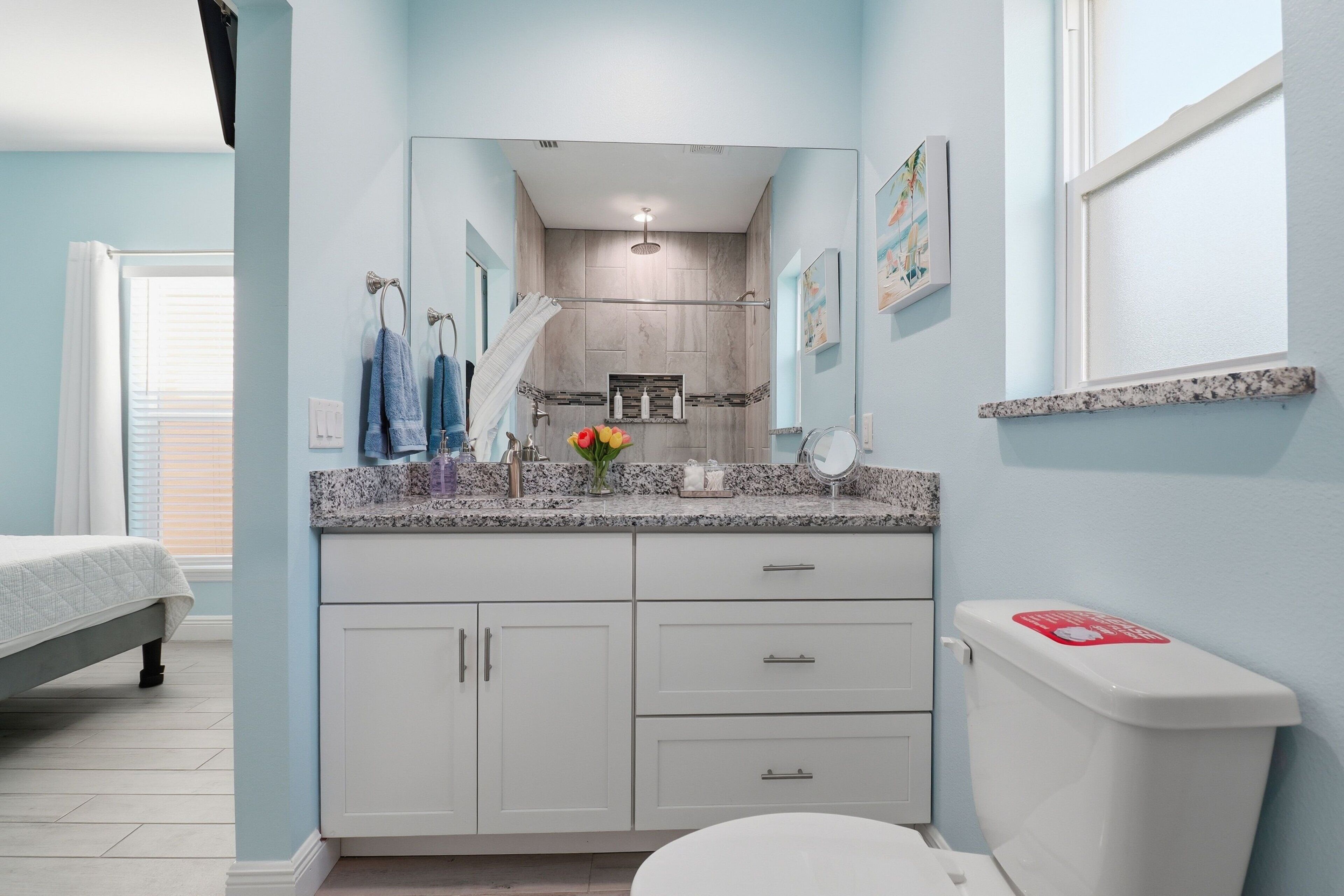 A bathroom with a shower, toilet, and sink, with a bed visible in the adjacent room.