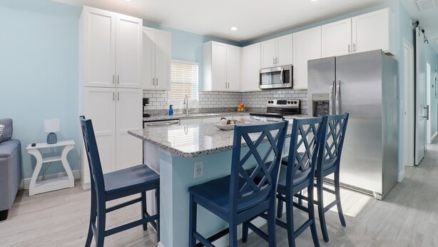 A modern kitchen with white cabinets, a central island with blue chairs, and stainless steel appliances. - New! Charming Lilac Cottage 111 (Punta Gorda)