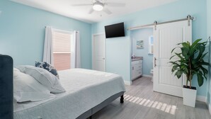 A bedroom with a bed, a ceiling fan, a TV, a door, a plant, and a bathroom with a sink and mirror. - New! Charming Lilac Cottage 111 (Punta Gorda)