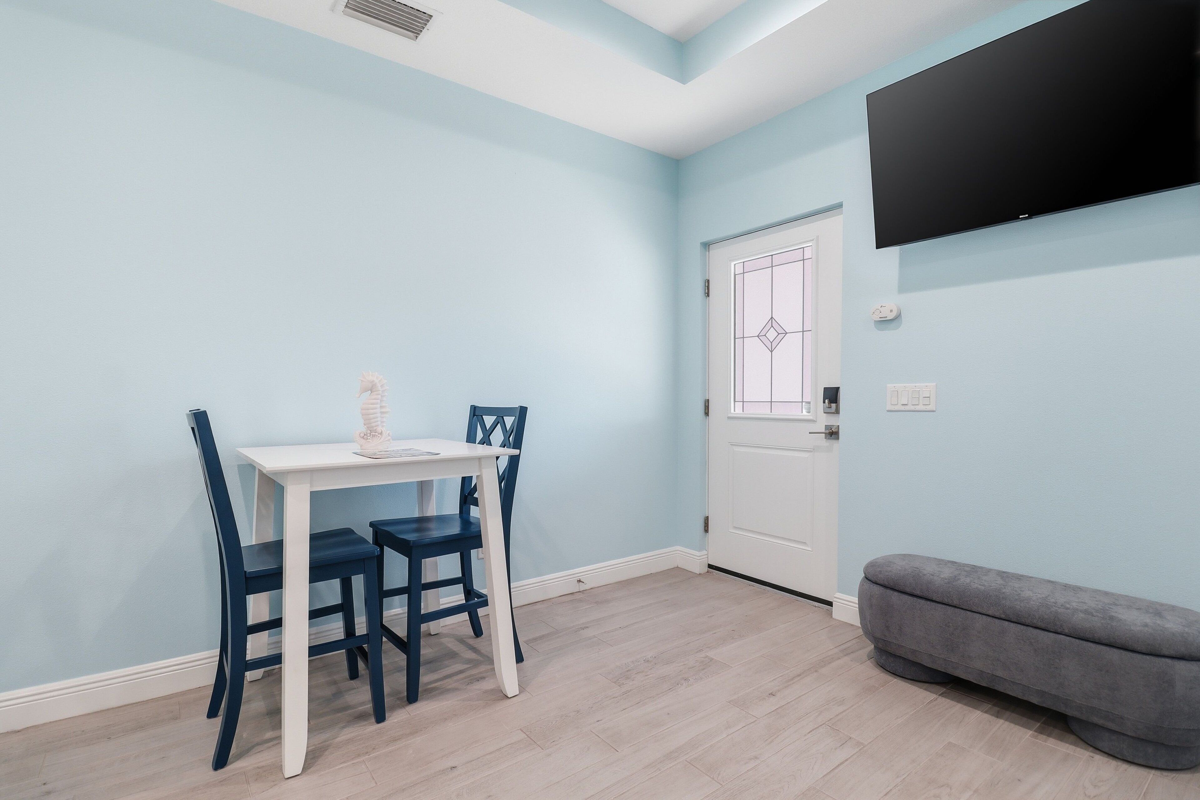 Een kamer met een witte tafel en blauwe stoelen, een grijze bank en een deur met decoratieve panelen, plus een tv aan de muur.
