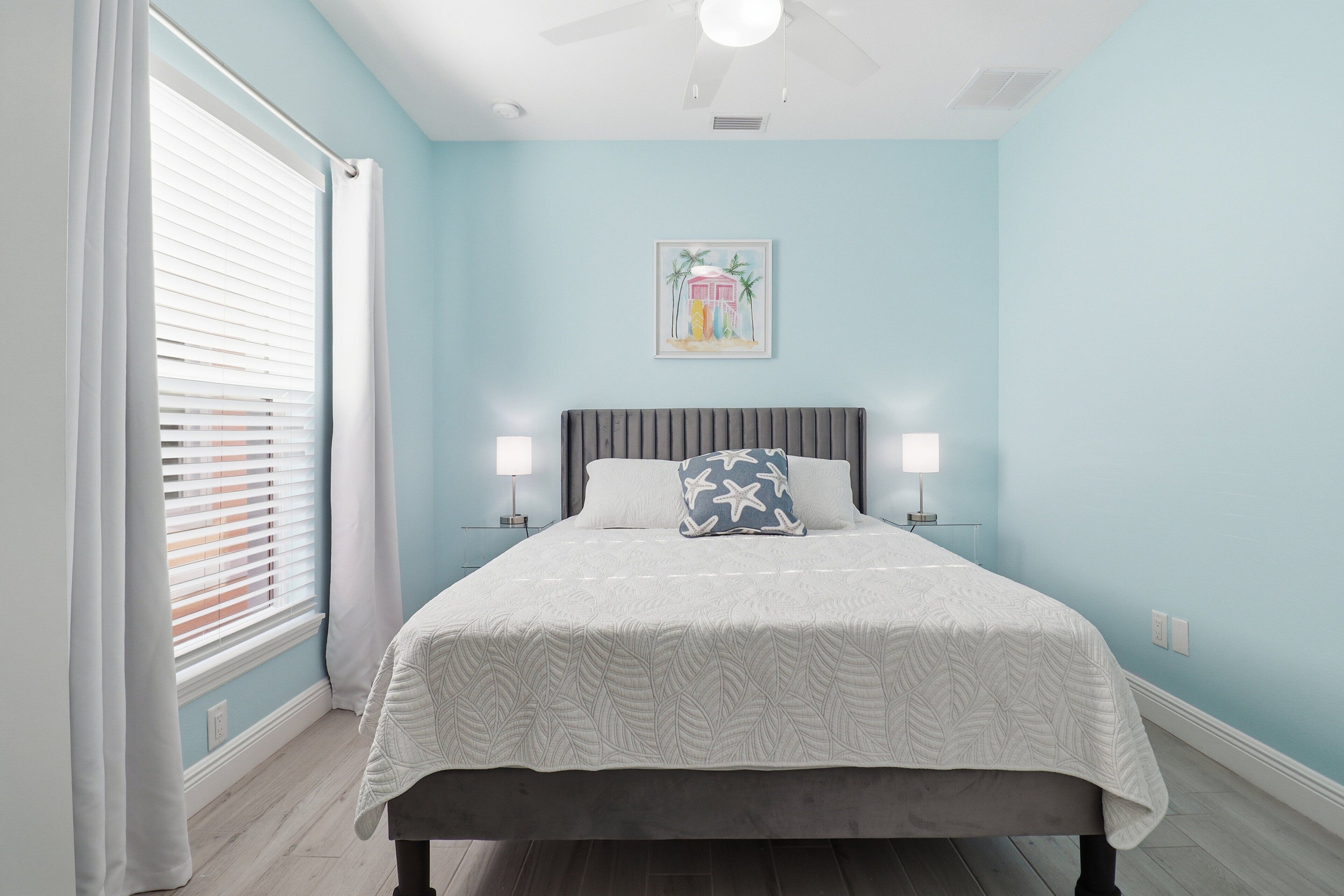 Une chambre à coucher avec un lit, un ventilateur de plafond, une fenêtre avec des stores et un tableau accroché au mur.