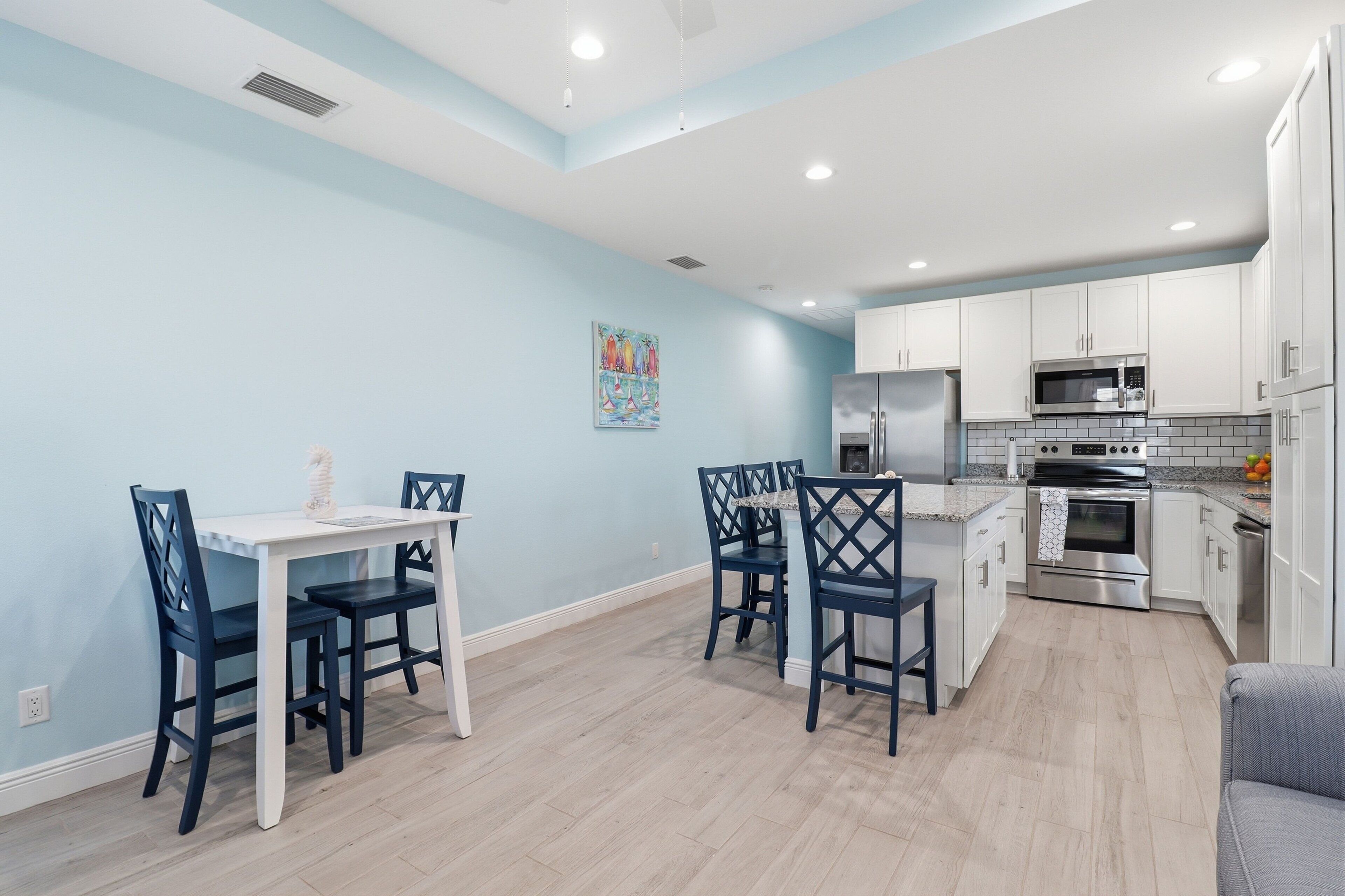 A modern kitchen with white cabinets, stainless steel appliances, and a dining area with blue chairs and a white table.