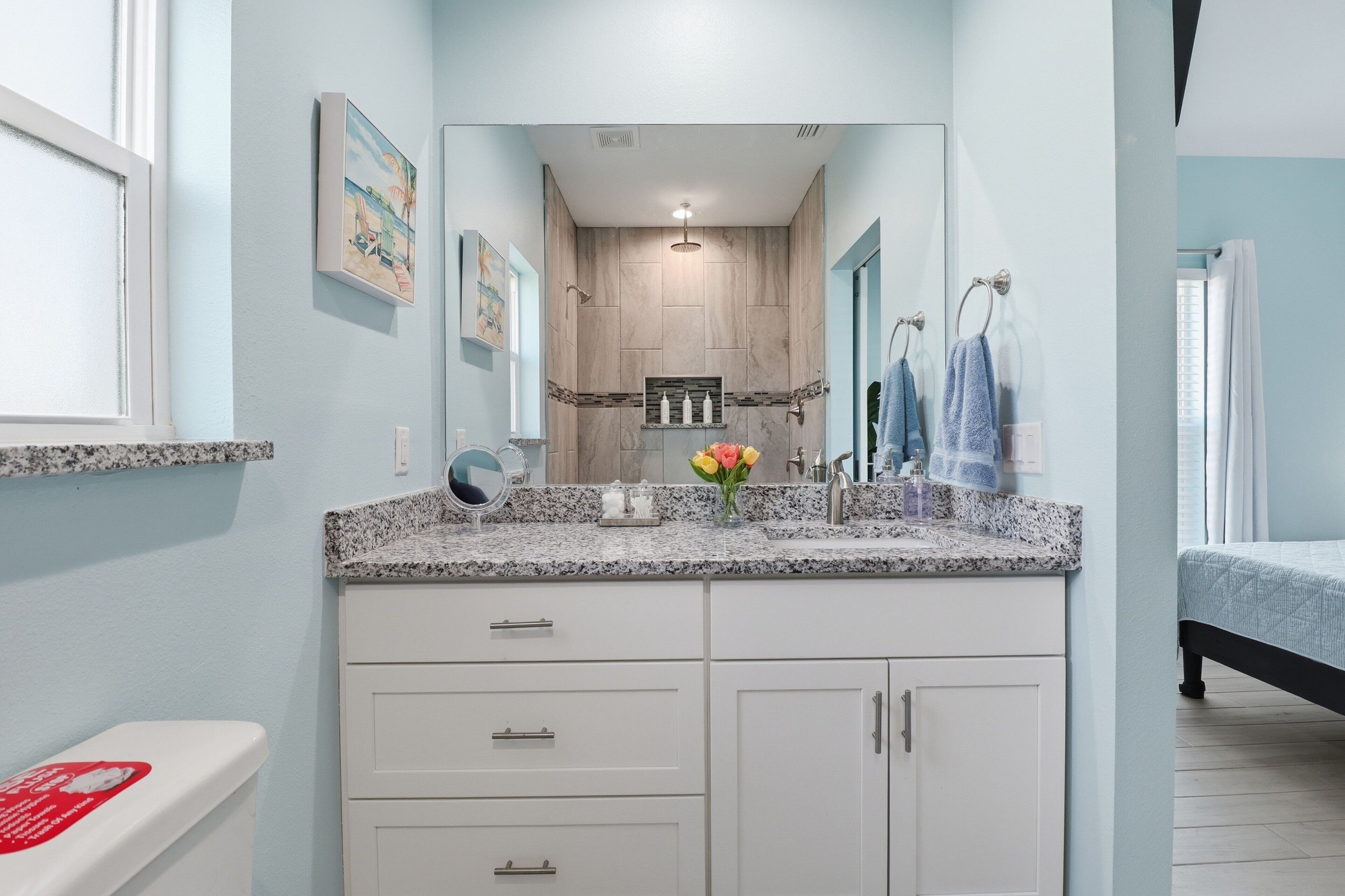 Une salle de bain avec un comptoir en granit, des armoires blanches, un miroir, une douche et un lit recouvert d’une courtepointe bleue.