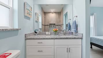 Une salle de bain avec un comptoir en granit, des armoires blanches, un miroir, une douche et un lit recouvert d’une courtepointe bleue.