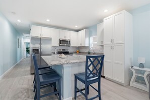 A modern kitchen with white cabinets, a stainless steel refrigerator, a microwave, and a granite countertop. There are two blue chairs and a small island with a sink. - New! Lovely Lilac Cottage 112 (Punta Gorda)