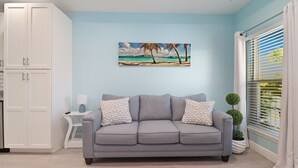 Ein Wohnzimmer mit grauem Sofa, einem Strandbild, einem weißen Schrank und einem Fenster mit Jalousien.