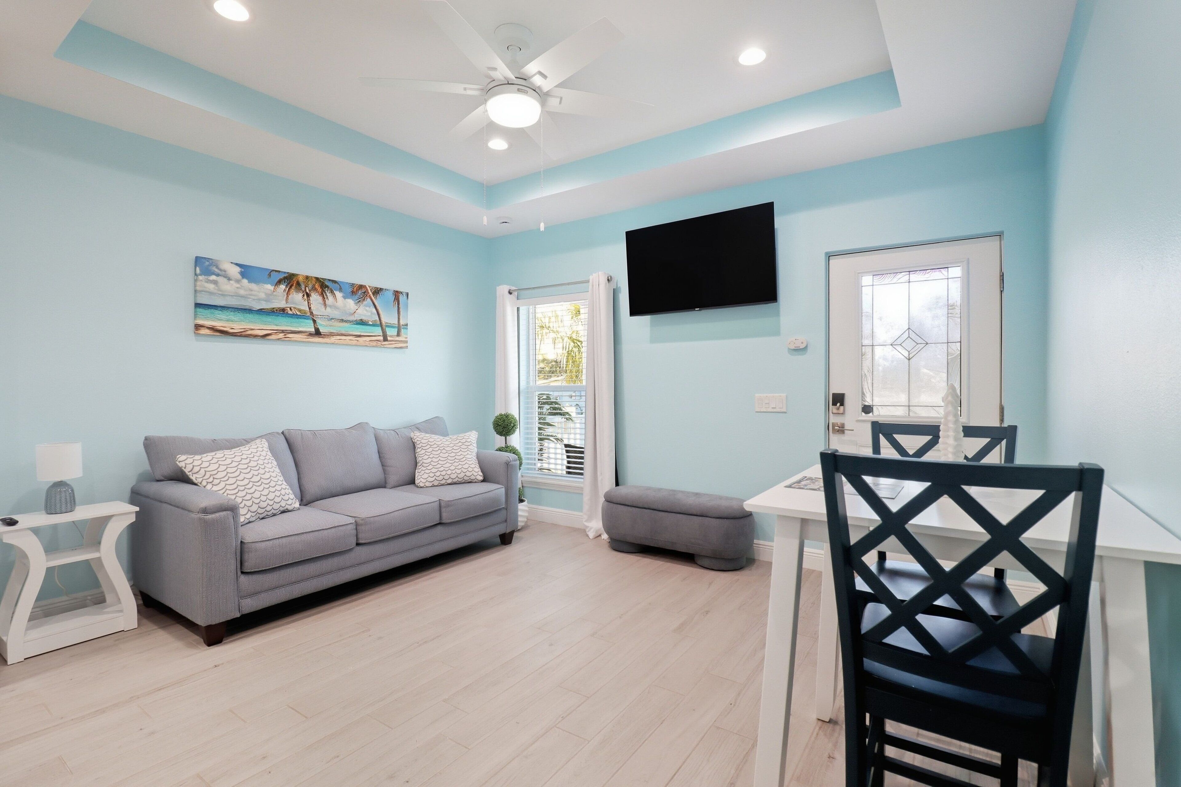 A living room with a grey sofa, a small table, a TV mounted on the wall, and a ceiling fan.