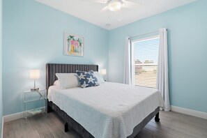 A bedroom with a bed, a nightstand, a ceiling fan, a window with blinds, and a wall painting. - New! Lovely Lilac Cottage 112 (Punta Gorda)