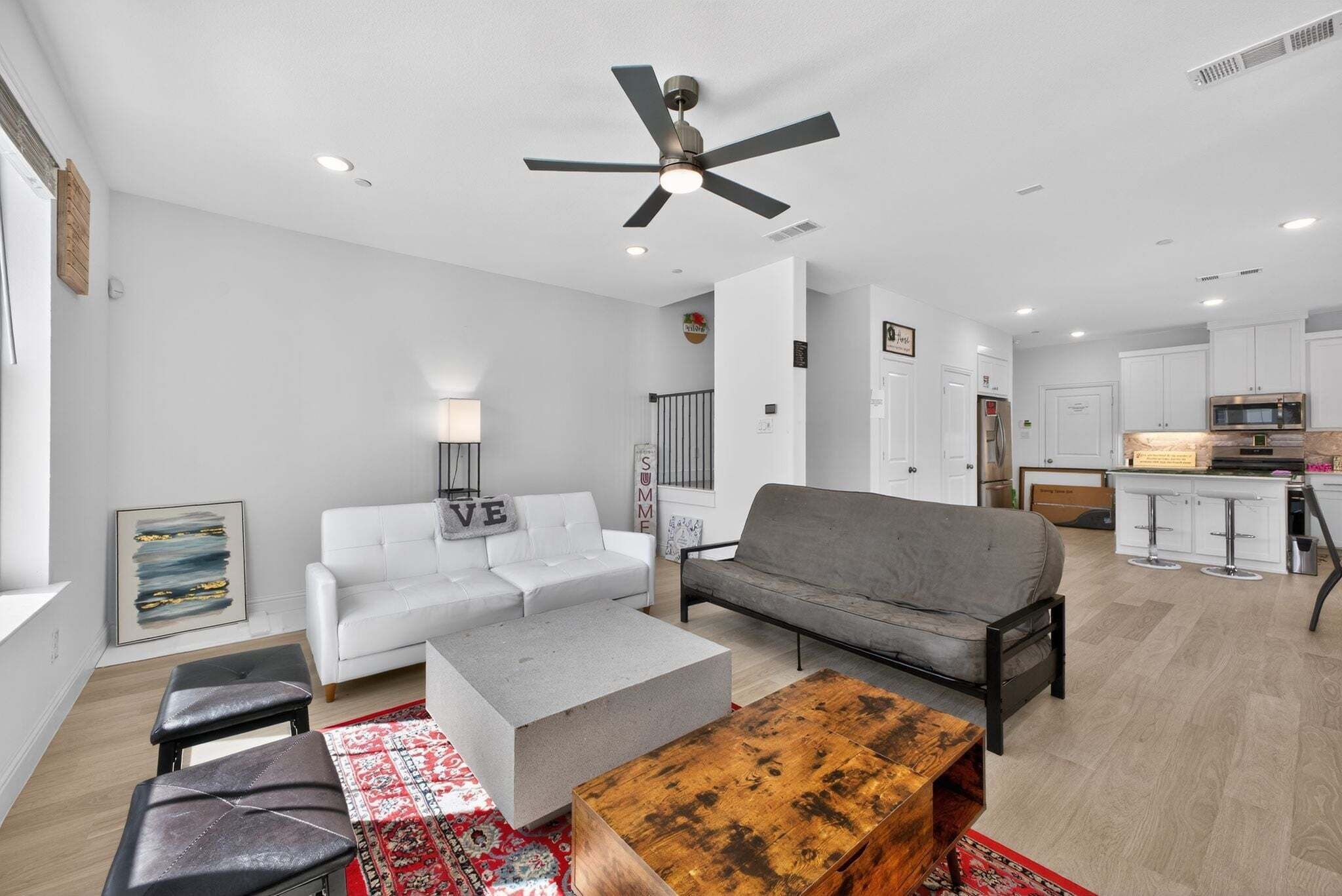 A modern living room with a white sofa, grey sofa, and a coffee table. There is a ceiling fan, a kitchen in the background, and a red patterned rug.