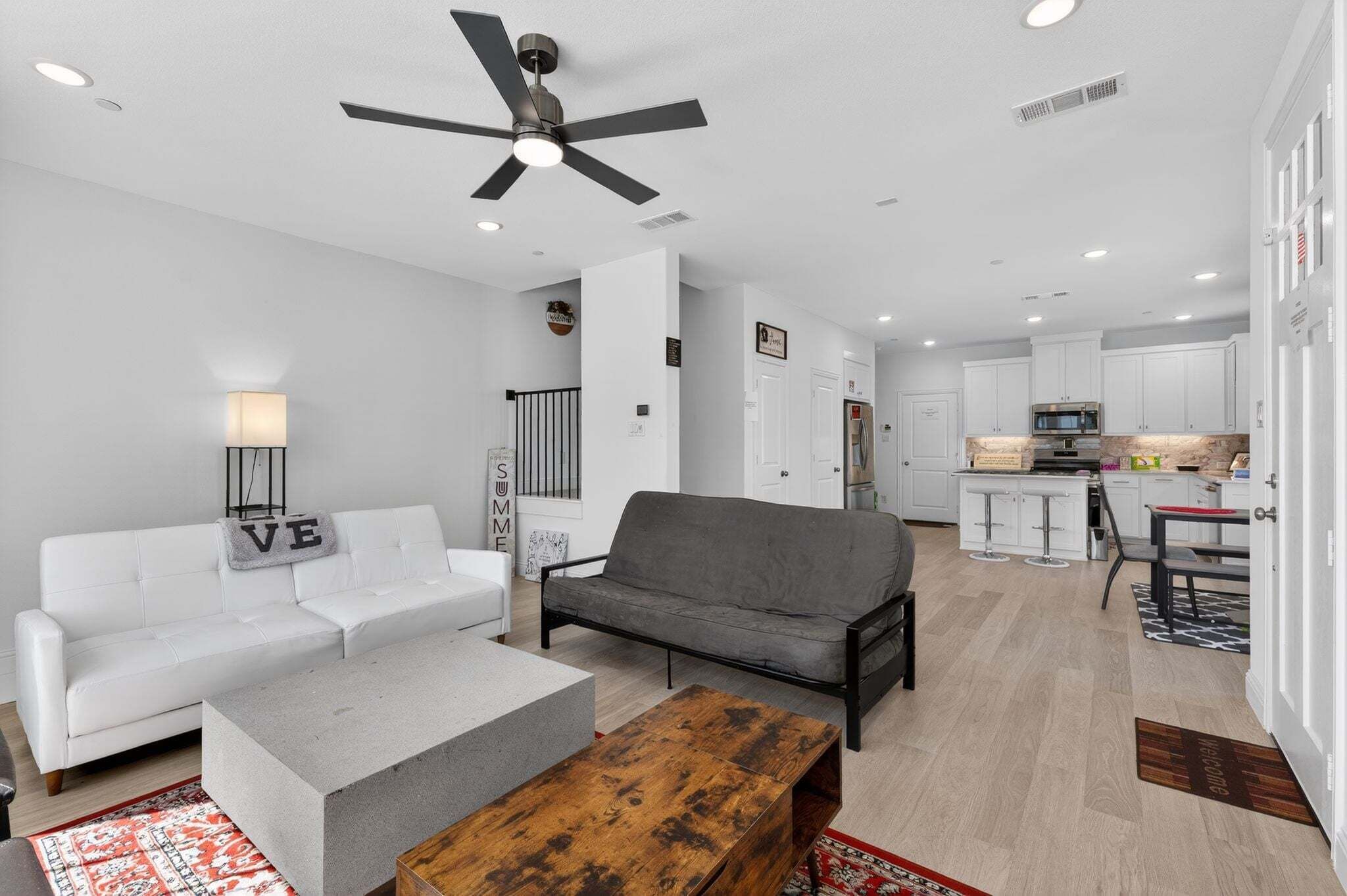 A modern living room with a white sofa, a grey sofa, a concrete coffee table, and a wooden table. There is a ceiling fan, recessed lighting, and a kitchen in the background.