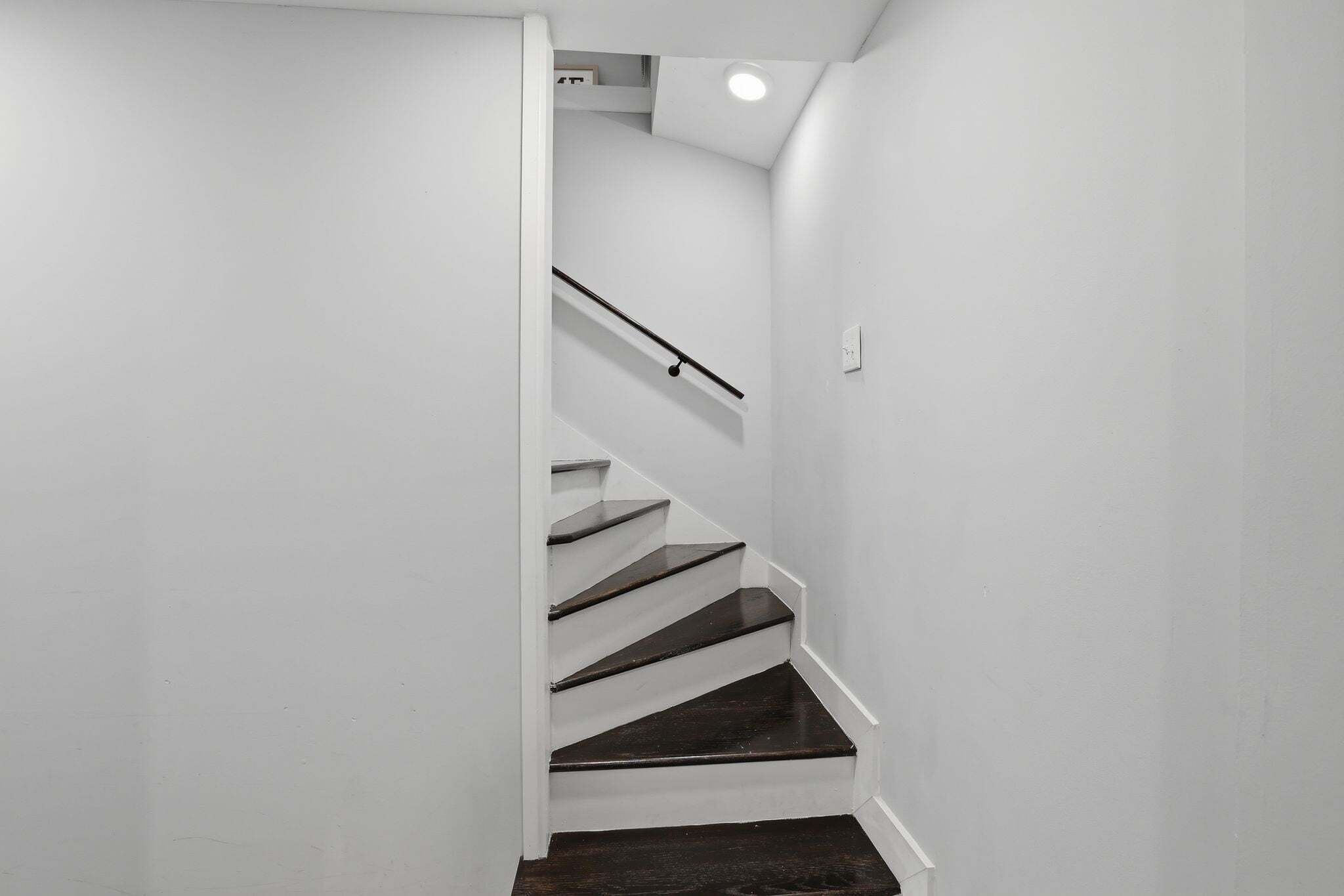 Un escalier aux murs blancs et aux marches sombres, menant à un palier où se trouve un luminaire.
