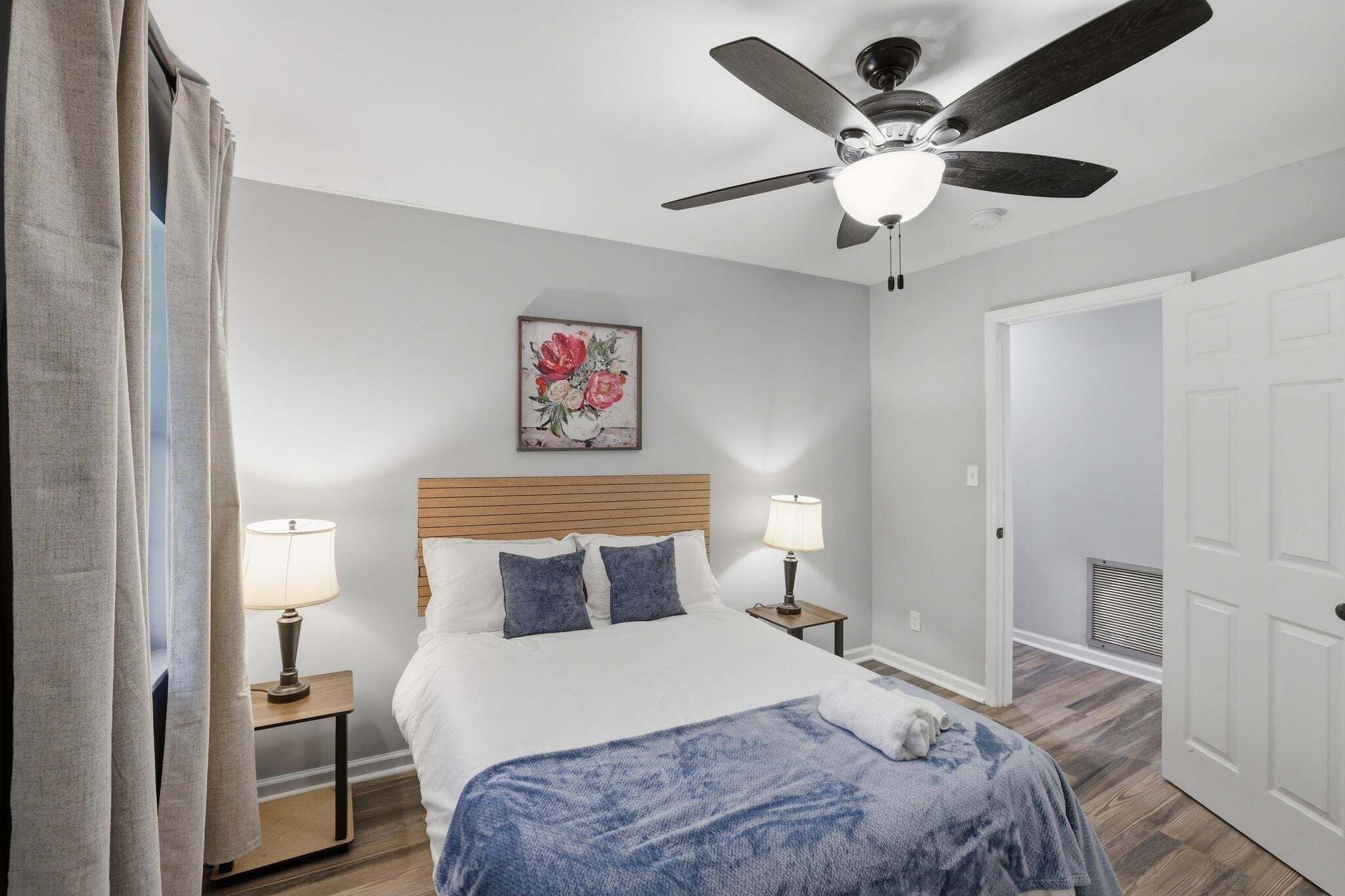 A bedroom with a bed, two bedside lamps, a ceiling fan, a door, and a wall-mounted picture.