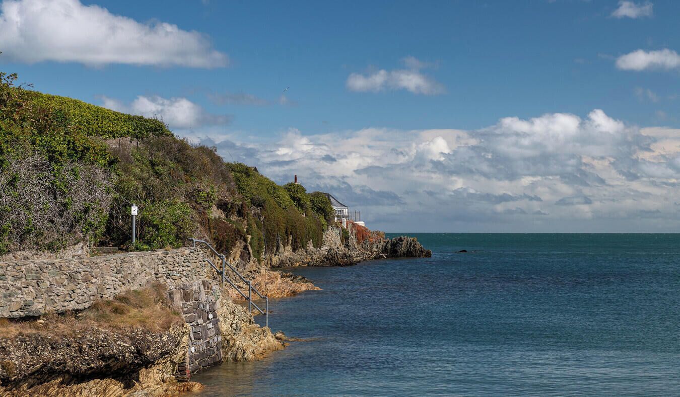 A coastal landscape with a stone wall, a building with a white roof, and a clear view of the sea.