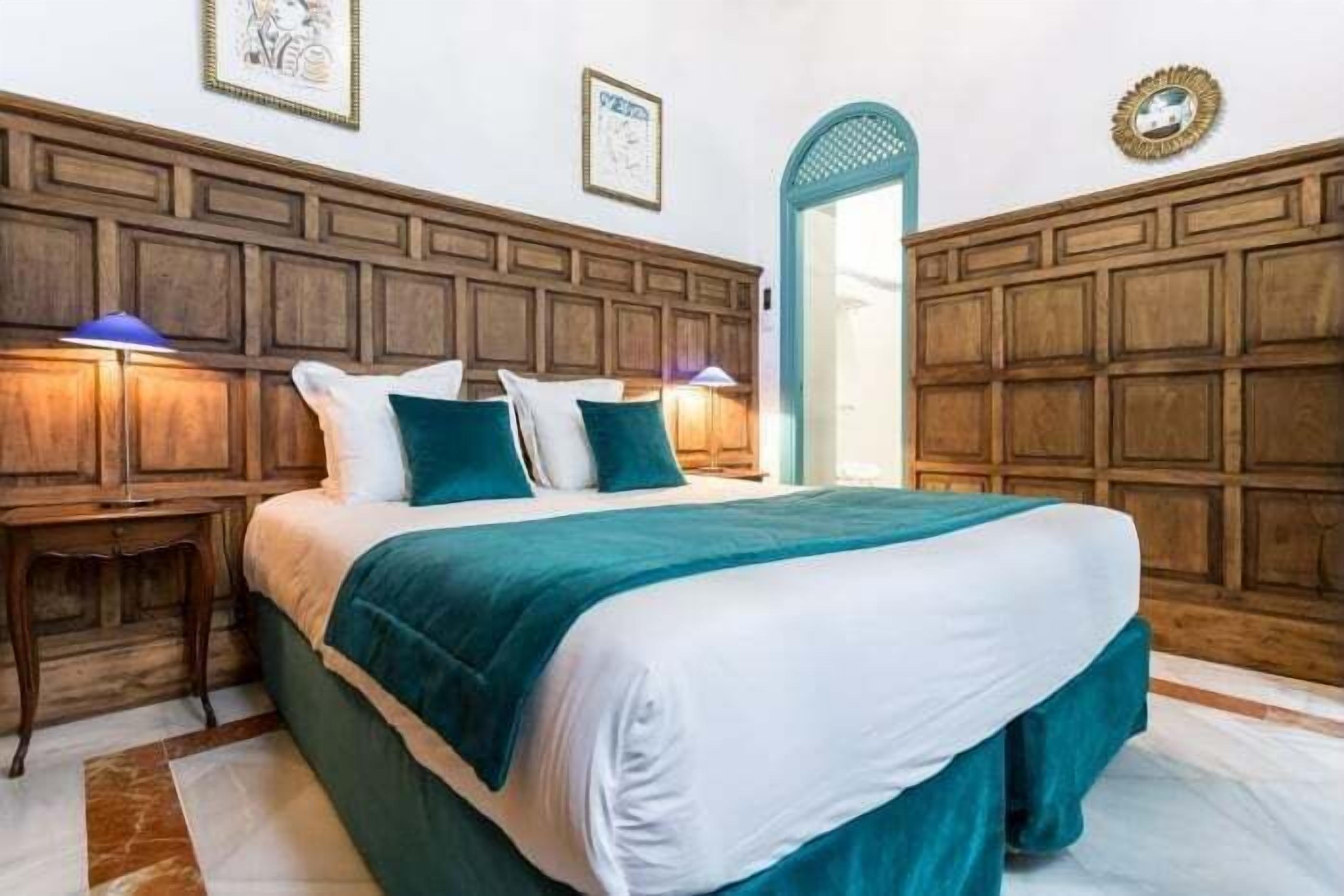 Un dormitorio con cabecera de madera, cama con ropa de cama blanca y turquesa, dos lámparas de noche y una mesita.