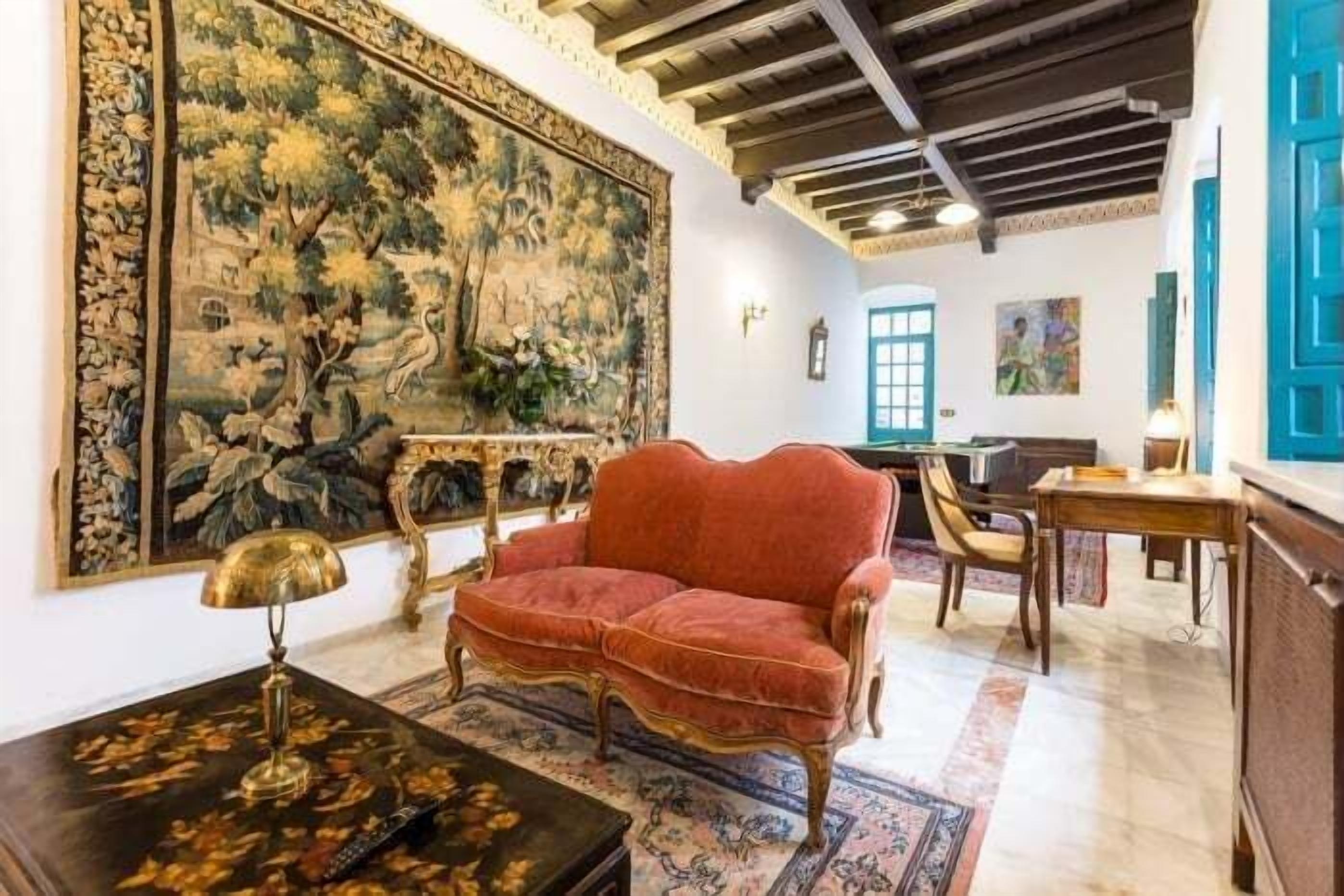 A room with a red velvet sofa, a large tapestry, a wooden table, and a blue door.