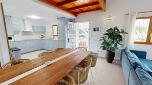 Uma cozinha moderna com armários azul-claros, uma mesa de jantar de madeira com cadeiras de vime, um sofá azul e uma grande planta em vaso.