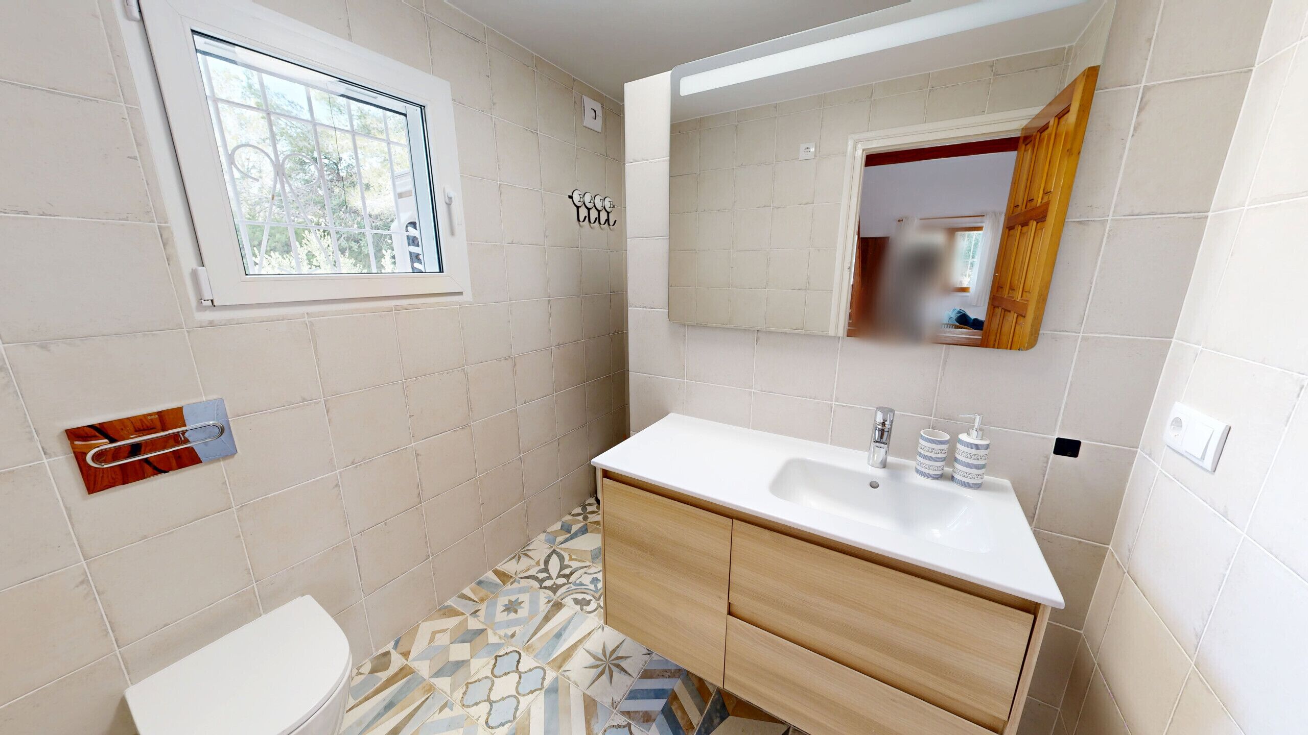 Un bany amb un lavabo blanc, un tocador de fusta, un vàter, una finestra i un mirall.