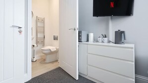 Et badeværelse med toilet, håndklædetørre og brusebad. Der er et hvidt skab med et fladskærms-tv monteret over det.