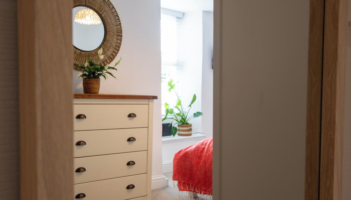 Un corridoio con una porta in legno, un comò beige con cassetti, uno specchio e una finestra con una pianta.