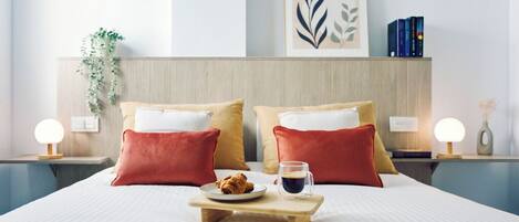 Een netjes opgemaakt bed met rode en beige kussens, een houten dienblad met een croissant en een kop koffie, en een ingelijst kunstwerk erboven.