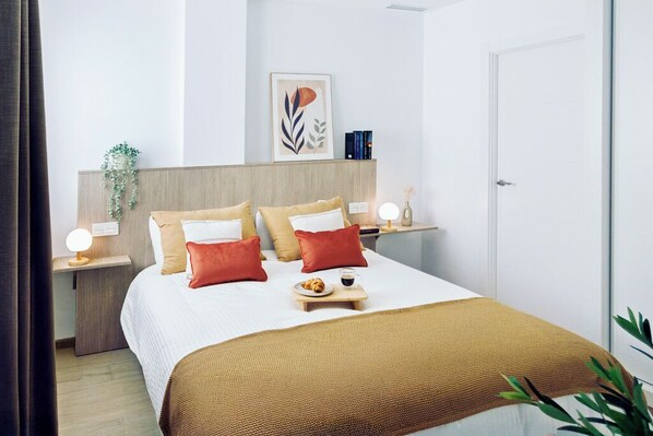 Une chambre d’hôtel bien rangée, avec un lit, des tables de chevet, une étagère fixée au mur, une porte et une plante.