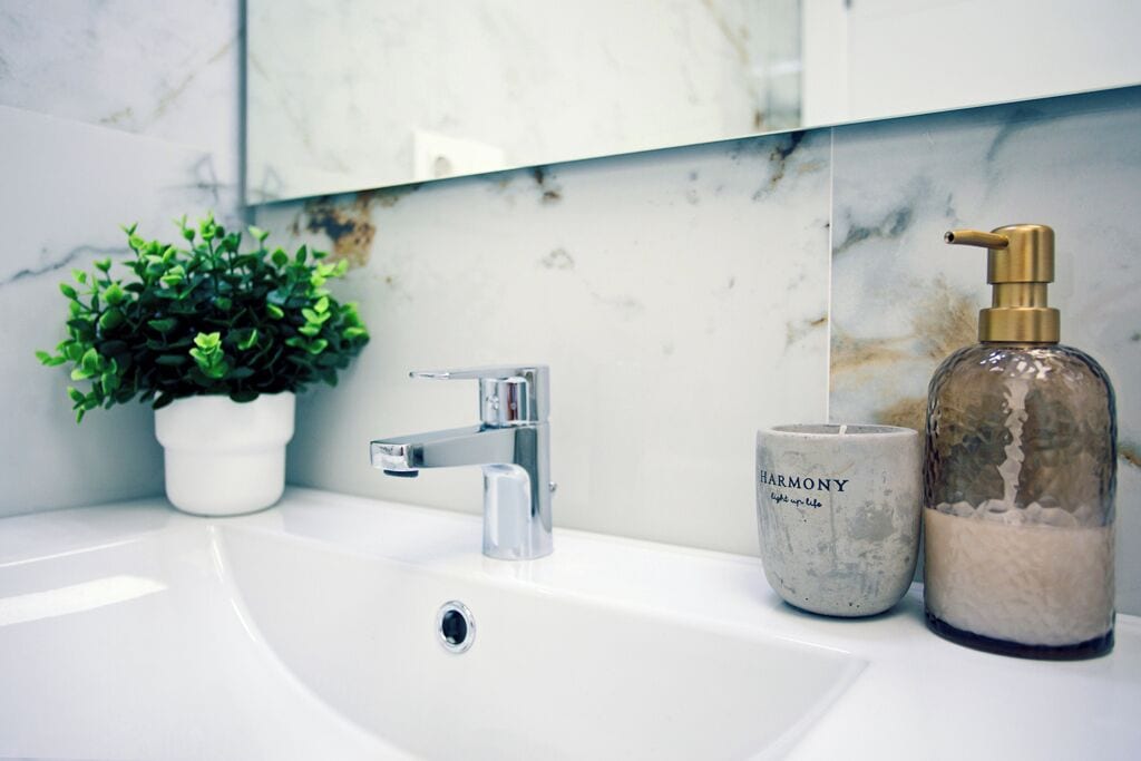 Un bagno con un lavandino bianco, un rubinetto cromato, una pianta in vaso e due candele etichettate 'HARMONY'.