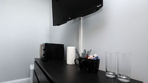 Un bureau noir avec un téléviseur à écran plat, un boîtier noir, une tasse noire contenant des stylos et deux verres transparents.