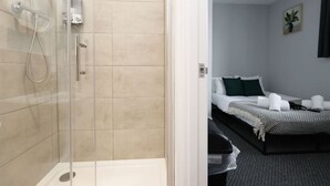 Hotellihuone, jossa on lasiseinäinen suihkutila, valkoisilla lakanoiden ja kuviollisella päiväpeitteellä varustettu sänky sekä pieni ruukkukasvi seinällä.