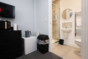 A modern bathroom with a shower, sink, and toilet. There is a black trash bin, a white coffee machine, and a black kettle on a black cabinet. A flat-screen TV is mounted on the wall.